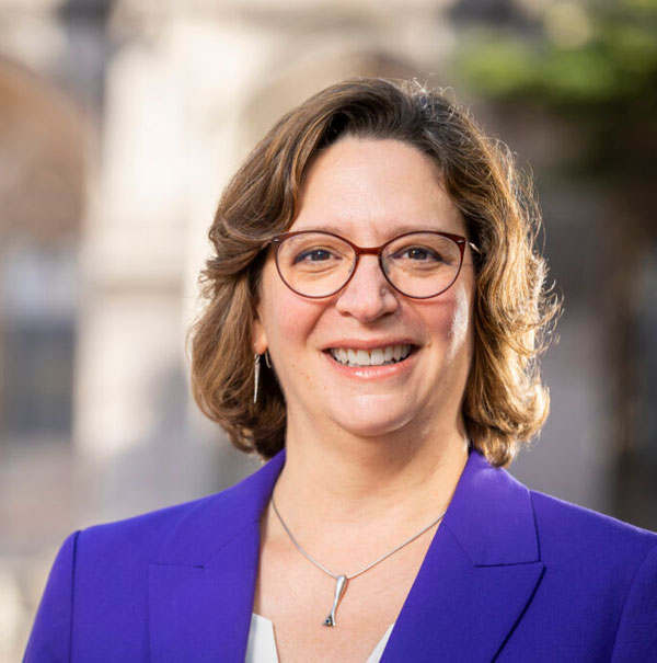 A headshot of Tricia Serio, a woman with chin-length brown hair wearing a purple blazer