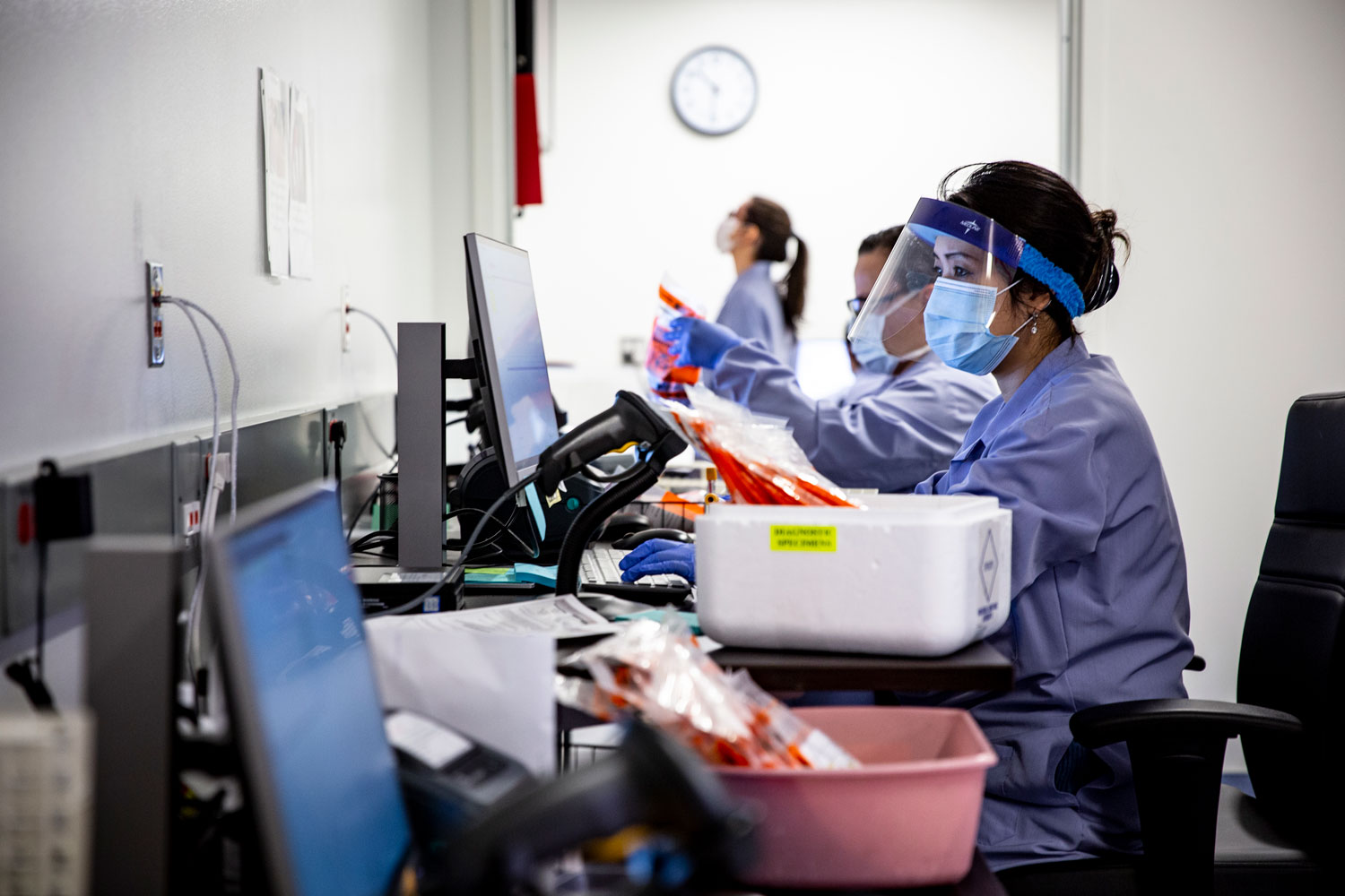 Three women in labcoats and face masks examine vials and computer screens.
