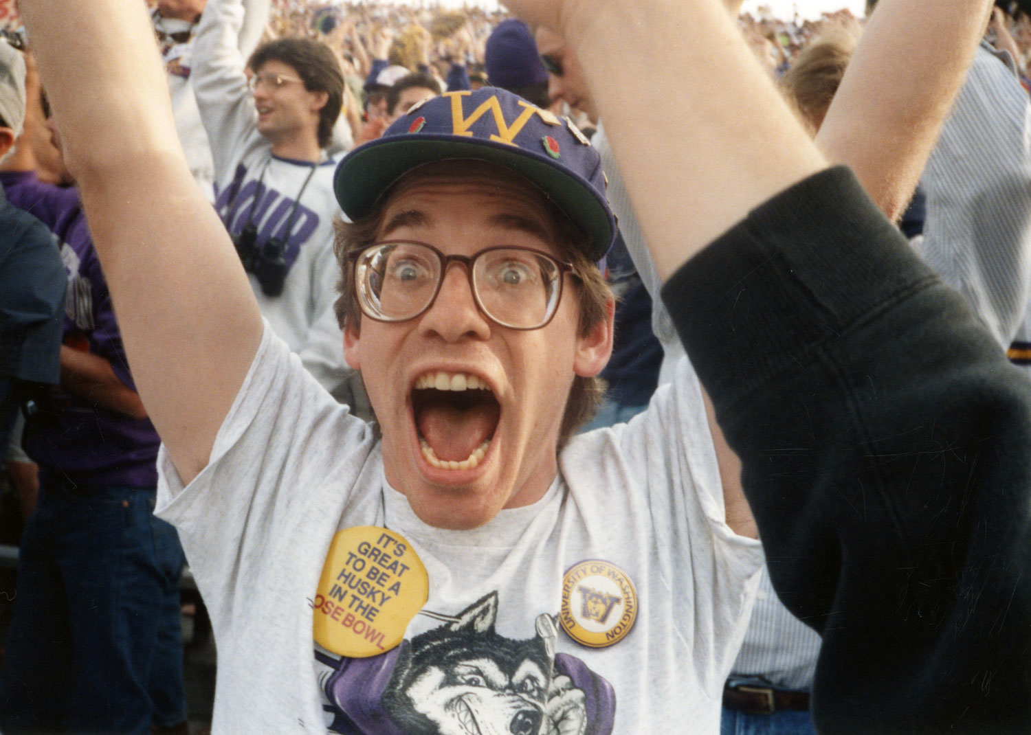 A man wearing glasses and Husky gear yells excitedly with his arms raised in a crowd.