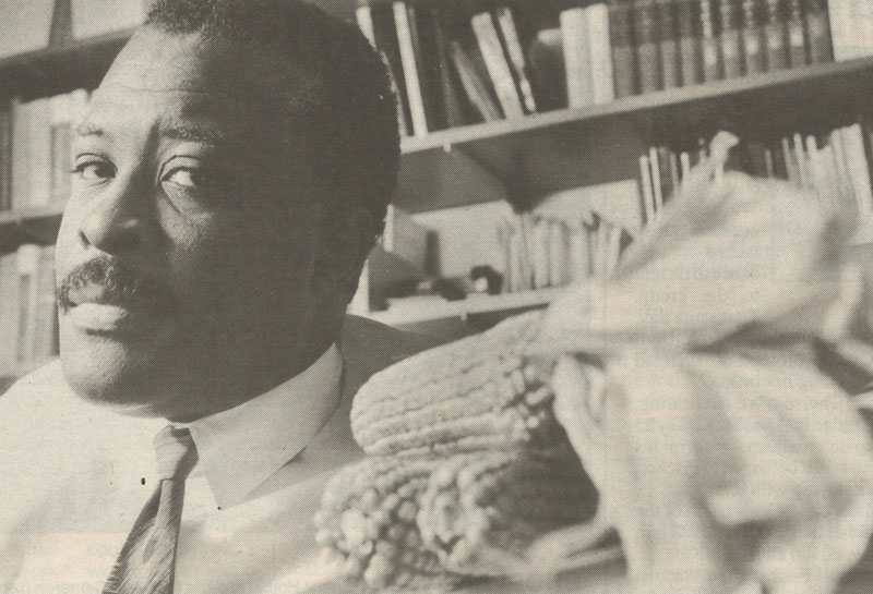 An old film photo of a man in a tie in a library next to a pile of corn stalks.