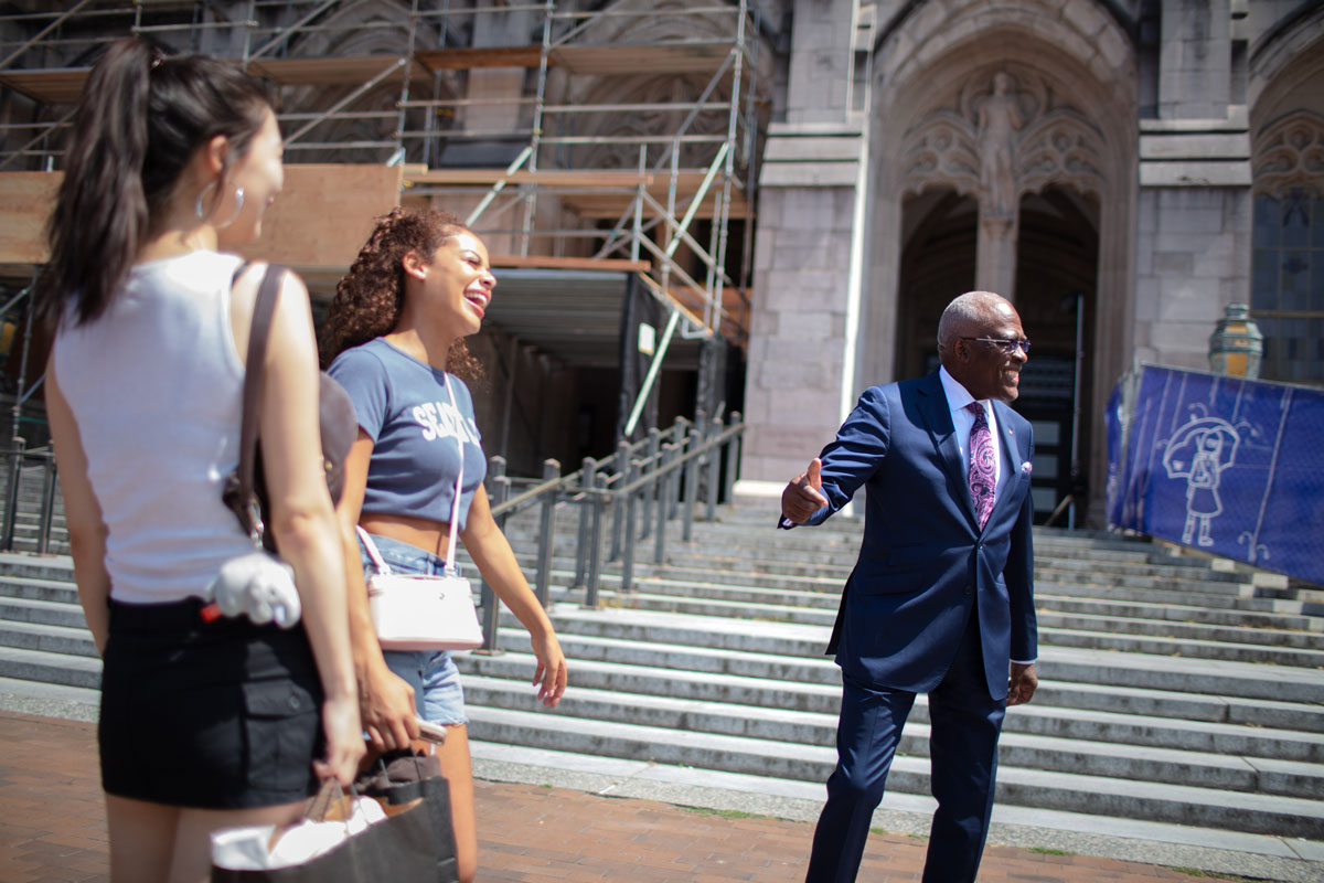 A man in a suit chats with two students on a college campus.