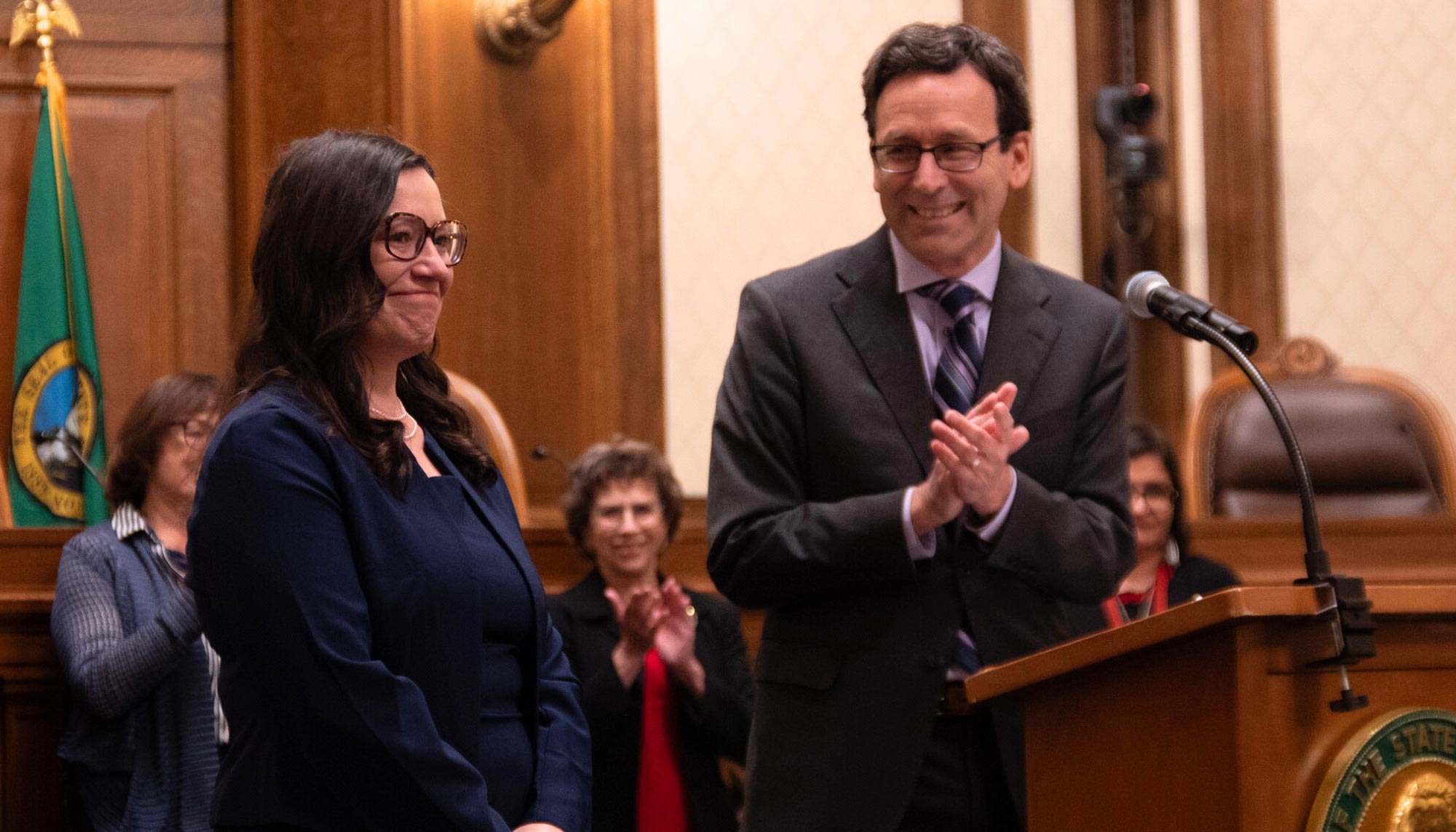 In a courtroom, Governor Bob Ferguson applauds at a podium next to Colleen Melody.