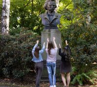 Three people gesturing to a statue of Edvard Grieg without a shiny nose