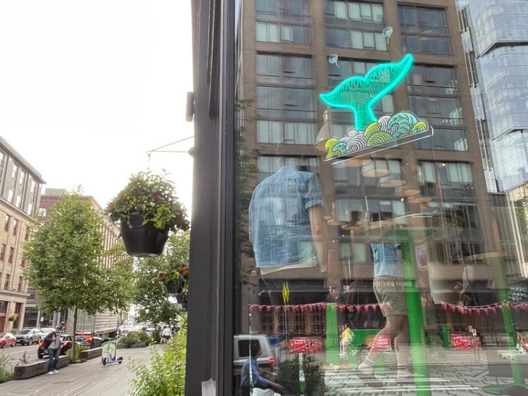 A neon-list whale tail sculpture hangs on a large window at Lowlander; people can be seen socializing inside